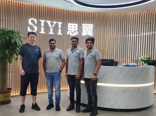 Four individuals are standing in an office lobby with "SIYI" displayed in large letters on the wall behind them. Three wear matching gray polo shirts, while one wears a dark blue t-shirt and black shorts, reflecting the streamlined efficiency of integrated supply chain management at Jane.