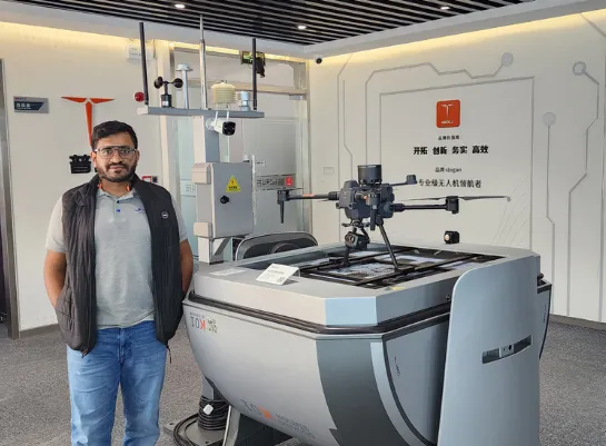 A man stands beside a large industrial drone in a modern showroom with technological equipment and signage on the walls, showcasing Jane's innovations in integrated supply chain management.