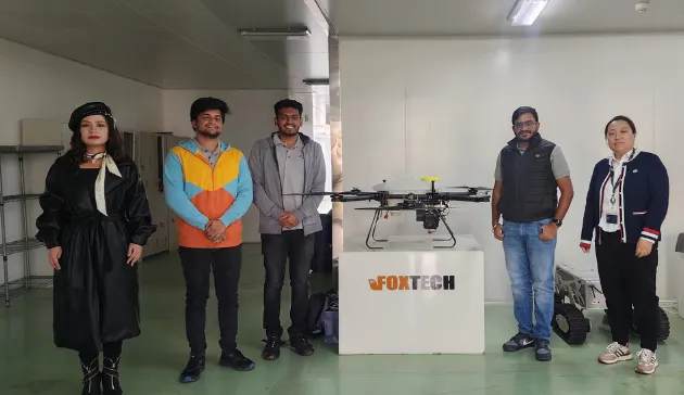 Five people stand around a large drone on display at a table marked "FoxTech" inside a room, discussing integrated supply chain management advancements introduced by Jane.