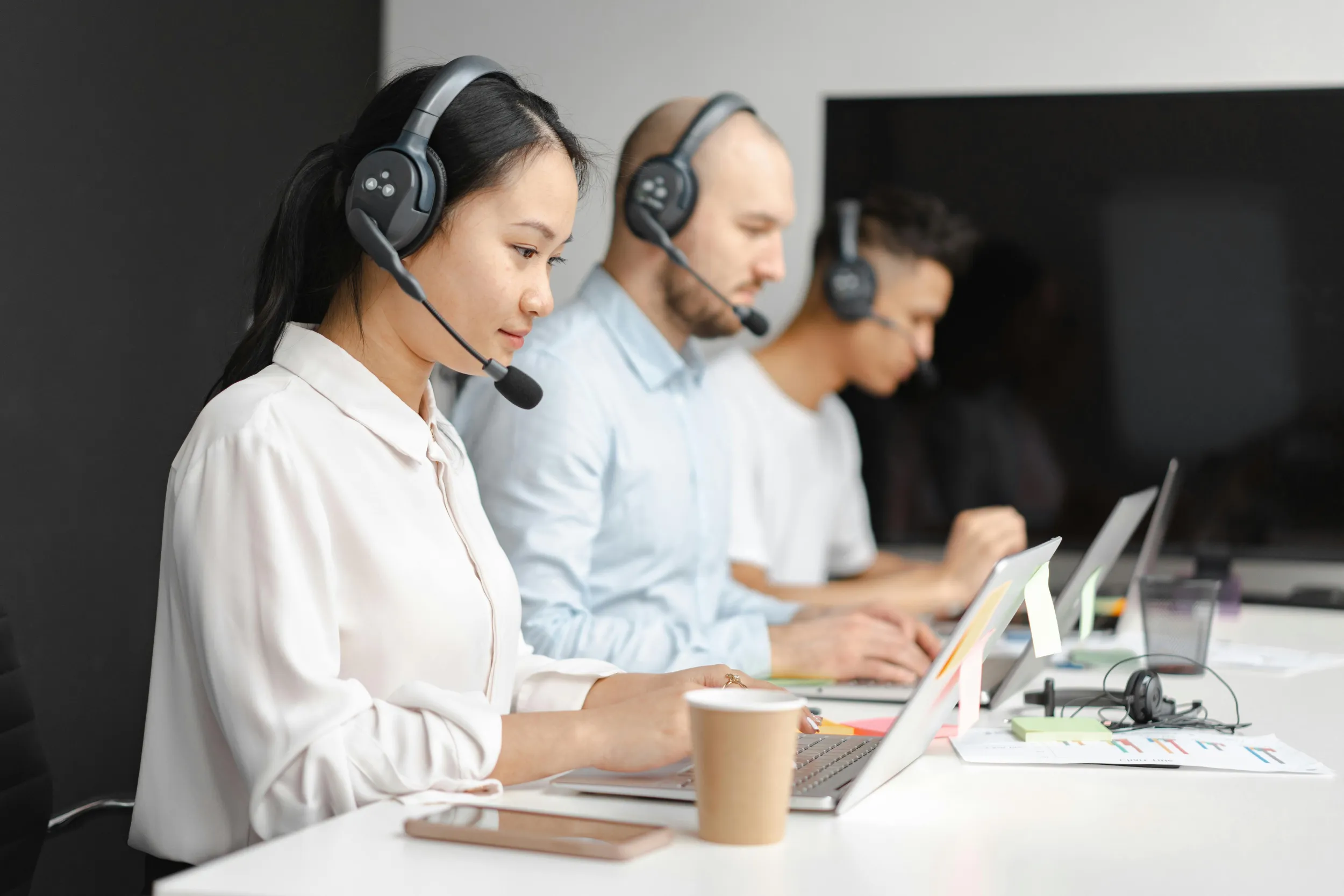 Three call center agents wearing headsets work at their laptops in a modern office, focusing on their tasks for Jane. A cup of coffee and smartphone are on the desk, suggesting the integration of efficiency and technology akin to seamless supply chain management.